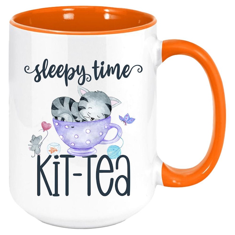 May include: White ceramic mug with an orange interior and handle. The mug features a whimsical design with a sleeping gray cat in a teacup, a mouse with a heart balloon, and the words "sleepy time" and "kit-tea".