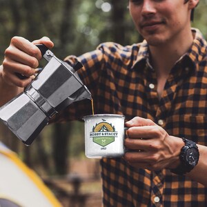 May include: A person pouring coffee from a silver stovetop espresso maker into a white enamel camping mug. The mug has a logo that reads "BOBBY & STACEY" with a mountain and tent design. The person is wearing a plaid shirt.