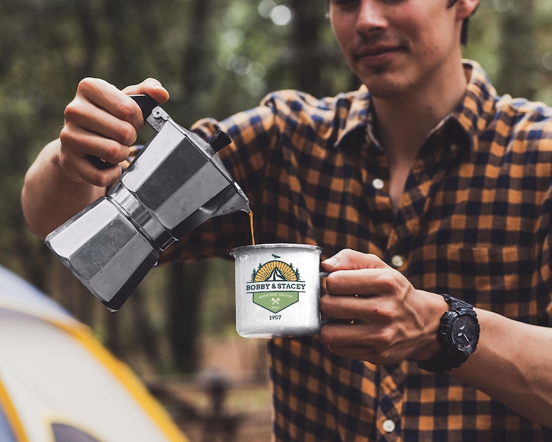 May include: A person pouring coffee from a silver stovetop espresso maker into a white enamel camping mug. The mug has a logo that reads "BOBBY & STACEY" with a mountain and tent design. The person is wearing a plaid shirt.