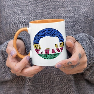 May include: White ceramic mug with an orange rim. The mug features a colourful graphic of an elephant silhouette inside a circle with a zebra print background.