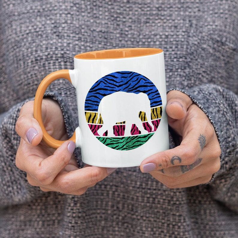 May include: White ceramic mug with an orange rim. The mug features a colourful graphic of an elephant silhouette inside a circle with a zebra print background.