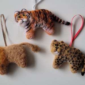 May include: Three felted animal ornaments, a lion, a tiger, and a leopard, each with a ribbon loop for hanging.