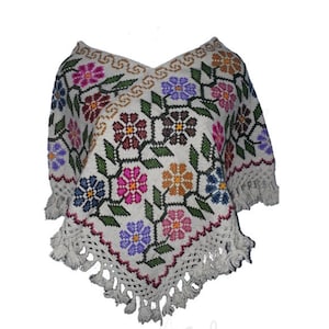 May include: Cream-coloured poncho with a V-neck and fringe. The poncho features a floral pattern in various colours, including pink, purple, blue, and orange. The edges are trimmed with a crocheted border and fringe.