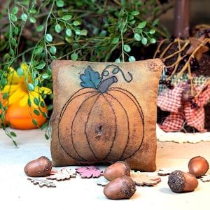 May include: A square fabric pillow featuring a hand-drawn pumpkin illustration with a teal leaf and brown stem. The pillow has a distressed, aged texture in shades of tan and brown. It is surrounded by several brown acorns and small leaf cutouts.