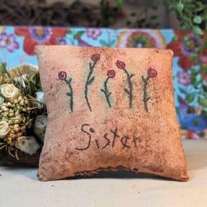 May include: A small, square, textured pillow with a light brown hue. The pillow features hand-drawn red roses on green stems and the word "Sister" in black script. The pillow is a decorative item.