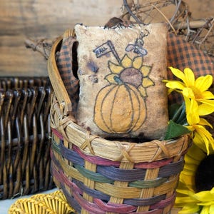 May include: A square fabric pillow with a distressed tan finish featuring a hand-drawn illustration of a pumpkin, a sunflower, and a small flag labeled FALL. The pillow is nestled inside a woven basket with multicolored splint accents.