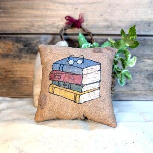 May include: A small, square, burlap pillow with a stack of books painted on the front. The books are blue, red, and yellow, with small glasses on top. The pillow is a neutral tan color.