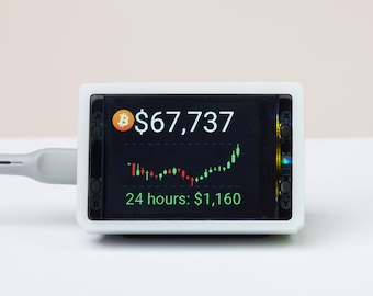 Puede incluir: Un dispositivo digital rectangular blanco de seguimiento de criptomonedas con una pantalla que muestra el logo de Bitcoin, el precio $67,737, un gráfico de velas rojo y verde, y el texto 24 hours: $1,160, conectado por un cable gris junto a un cubo de rompecabezas colorido.