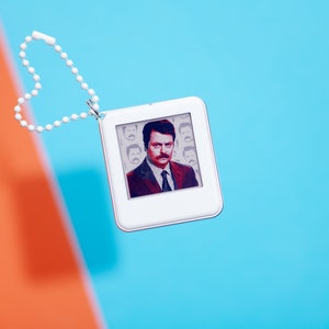 May include: A white keychain with a heart-shaped chain and a small square charm featuring a color image of a man with a mustache and a suit. The charm has a white border and a black background.