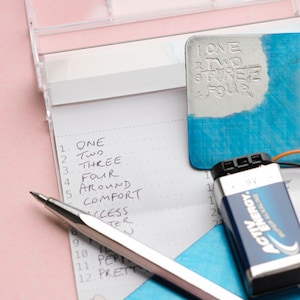 May include: A white notepad with a list of words numbered 1 through 12. The words are "ONE", "Two", "THREE", "FOUR", "AROUND", "COMFORT", "ACCESS", "TER", "PEP", and "PRETT". A silver pen is lying on top of the notepad. A blue piece of paper with the words "ONE", "TWO", "THREE", and "FOUR" written on it is in the background. A 9V battery is also in the background.
