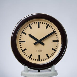 May include: A round, vintage clock with a dark brown frame and a cream-colored face. Black hands point to the time, and black dashes mark the minutes. The clock is supported by a light gray base. The text "RFT" is visible on the clock face.
