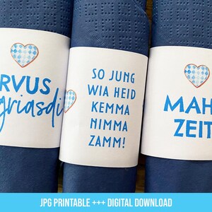 Könnte beinhalten: Drei blaue Servietten mit weißen Papierbändern. Die Bänder haben unterschiedliche Texte in Deutsch. Das erste Band sagt "Servus griasdi" mit einem blau-weißen Herzen. Das zweite Band sagt "So jung wia heid kemma nimma zamm!" Das dritte Band sagt "Mahl Zeit".