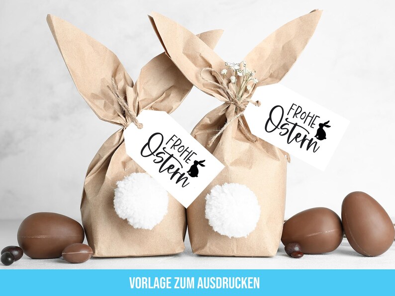 May include: Two brown paper bunny bags with white pom-poms and tags that say "Frohe Ostern" with a bunny silhouette. The bags are sitting on a white surface with chocolate Easter eggs.