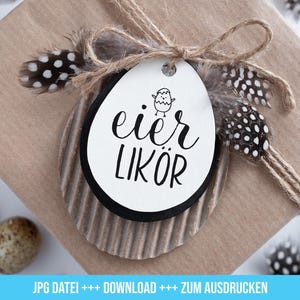 Op de afbeelding: Een wit cadeau-etiket met zwarte tekst die "eier likör" luidt en een kleine zwart-witte illustratie van een kuiken. Het etiket is vastgebonden met touw en versierd met bruine en witte veren.