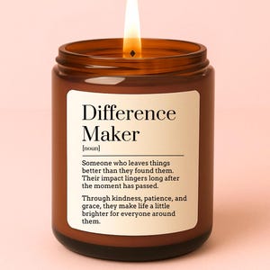 May include: A burning brown glass candle with a white label. The label reads "Difference Maker" and includes a definition. The candle's flame is bright yellow. The candle is in a glass jar.