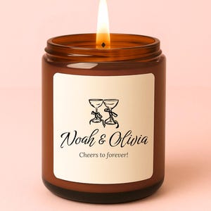 May include: A lit brown glass candle jar with a white label featuring the names "Noah & Olivia" and the phrase "Cheers to forever!" in elegant script. The label also includes a graphic of two champagne glasses.