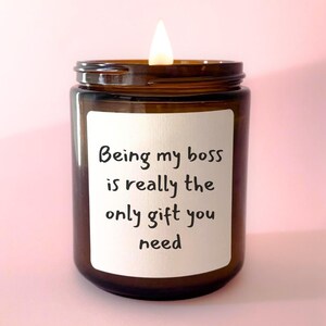 May include: A brown glass candle with a white label that reads "Being my boss is really the only gift you need". The candle is lit and the flame is visible.