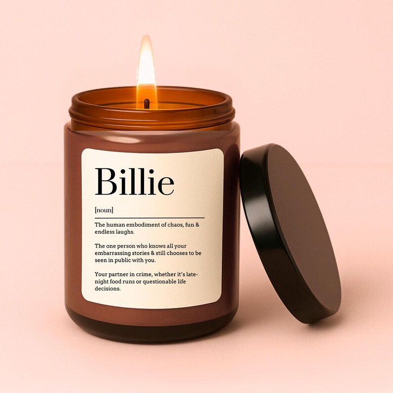 May include: A brown glass candle with a black lid. The candle has a white label with the word "Billie" and a definition of the word. The candle is lit and the flame is visible.