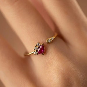 Può includere: Un anello color oro con una pietra preziosa rosso scuro a forma di pera e piccole pietre chiare. Il design aperto presenta una fascia divisa, con il gruppo di pietre preziose da un lato e due pietre più piccole dall'altro. L'anello è mostrato su una mano.