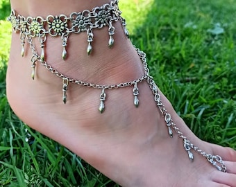 May include: Silver ankle bracelet with a delicate chain and dangling charms. The bracelet has a floral design and is perfect for layering.
