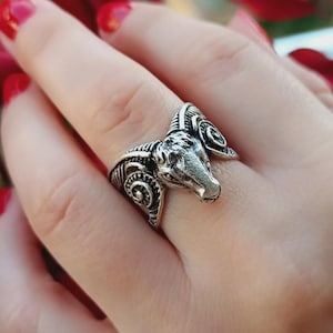 May include: Silver-toned ring featuring a ram's head design. The ring has detailed carvings and is worn on a finger. The ram's head is the focal point, with curved horns and a textured surface. The background is blurred, with red and green tones.