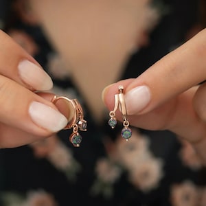 May include: Rose gold hoop earrings with dangling iridescent gemstone accents. The earrings are held up against a dark floral background, showcasing their delicate design and colorful sparkle. A close-up shot highlights the jewelry's craftsmanship.