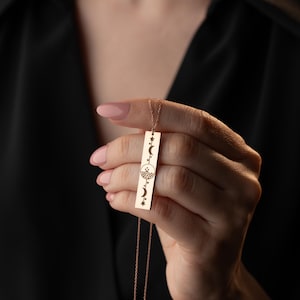 Puede incluir: Un collar de oro con un colgante rectangular largo que presenta un diseño de luna y estrellas. El colgante está grabado con una luna creciente, estrellas y un círculo con una montaña en su interior.