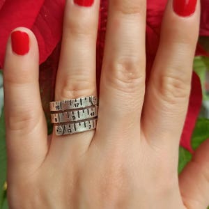 May include: A silver ring with three bands, each featuring a ruler design with numbers 16, 17, 18, 19, 2, and 9. The ring is worn on a finger with red nail polish.