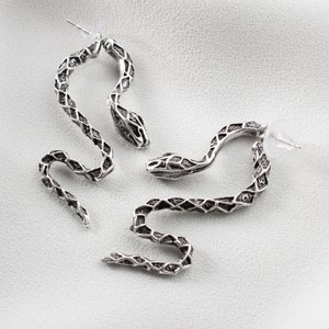 K&ouml;nnte beinhalten: Ein Paar silberne Schlangenohrringe. Die Ohrringe bestehen aus einem detaillierten Schlangendesign mit einer strukturierten Oberfl&auml;che. Die Ohrringe sind an einem Stift mit einem Schmetterlingsverschluss befestigt.