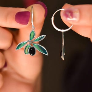 Könnte beinhalten: Ein Paar silberne Ohrringe mit einem grünen Olivenzweig-Design und einem schwarzen Oliven-Anhänger. Die Ohrringe sind baumelnd und haben ein Ringdesign.