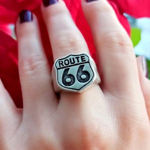 Può includere: Anello color argento con l'iconico design a scudo della Route 66. L'anello mostra le parole "ROUTE" sopra il numero "66" in grassetto e in nero. Un classico gioiello.