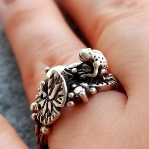 Puede incluir: Un anillo de plata con un diseño detallado que presenta una rana sentada en una hoja. El anillo se lleva en un dedo.