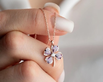 May include: A rose gold necklace featuring a four-leaf clover pendant. The clover is adorned with purple gemstones and small white stones, with a single white stone at the center. The necklace is held by a person.