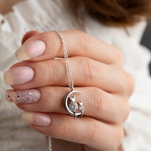 May include: A silver necklace with a crescent moon pendant featuring two small cats with sparkling stones for eyes.