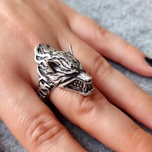 May include: A silver ring with a wolf head design. The wolf's mouth is open and its teeth are visible. The ring is on a person's finger.