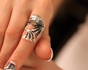 May include: A silver frog ring with detailed sculpting. The frog is sitting on a finger with a white nail polish.