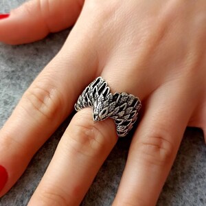 May include: A silver-toned ring in the shape of a snake, with detailed scales and a textured design. The ring is worn on a finger, showcasing its intricate craftsmanship and unique style. The ring's design features a snake head.