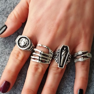 May include: Four silver rings on a hand. One ring features a skull, one ring features a skeleton hand, one ring features a coffin with a figure inside, and one ring features a face.