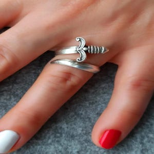 May include: A silver ring featuring a sword design. The ring is wrapped around the finger with the sword pointing upwards.