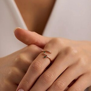 May include: A gold-colored ring with a branch and leaf design. The ring features two small green leaves and wraps around the finger. The ring is worn on a hand with a white background.