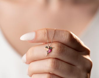May include: A gold-colored hummingbird pendant necklace. The hummingbird has a pink gemstone body, green gemstone wings, and is suspended from a delicate chain. The necklace is held up by a person's fingers.