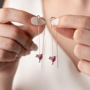 Può includere: Un paio di orecchini a colibrì in oro rosa. Ogni orecchino presenta un ciondolo a colibrì con pietre preziose rosa e verdi, sospeso a una delicata catena. Gli orecchini sono tenuti da un gancio curvo.