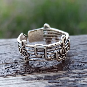May include: Silver-toned ring featuring musical notes and treble clef symbols. The ring's design incorporates musical staff lines and note shapes, creating a unique accessory for music lovers. The ring is set against a blurred background.