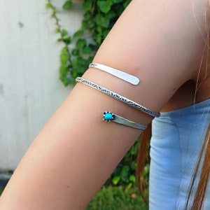 May include: Silver arm cuff with a turquoise stone accent. The cuff is textured and has a simple design.
