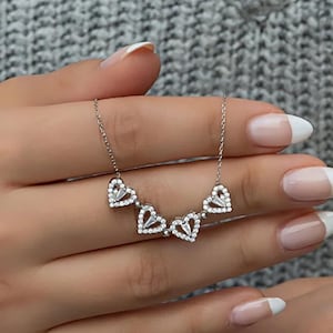 May include: A silver necklace featuring four heart-shaped pendants, each encrusted with small, clear gemstones. The hearts are connected by a delicate chain. The necklace is displayed against a soft gray background.