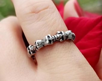 May include: A silver-colored ring featuring a repeating pattern of small skull designs. The ring is worn on a finger, with a red flower in the background. The skulls are detailed, giving the ring a gothic or edgy aesthetic.