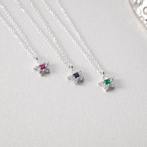 May include: Three silver necklaces with delicate flower pendants. Each pendant features a small, sparkling gemstone in the center: red, blue, and green.