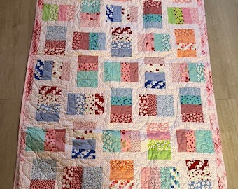 Puede incluir: Una colcha de retazos con fondo blanco y cuadrados de varios colores y patrones, incluyendo rojo, rosa, azul, verde y naranja. La colcha está bordeada con un ribete rosa.