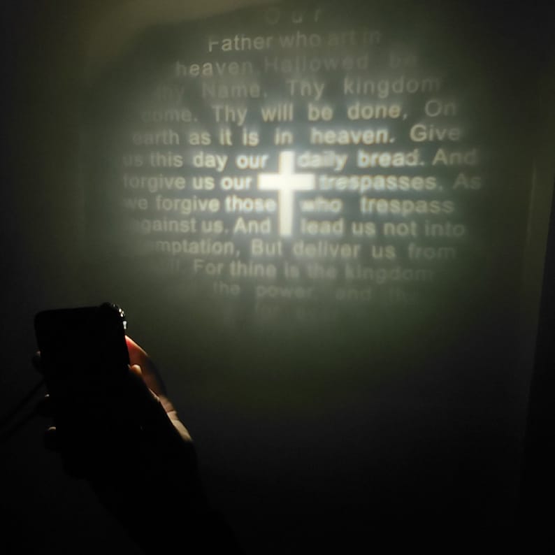 K&ouml;nnte beinhalten: Nahaufnahme eines Projektors, der das Vaterunser mit einem wei&szlig;en Kreuz in der Mitte anzeigt. Der Text ist leicht verschwommen. Eine Hand h&auml;lt ein schwarzes Smartphone vor dem Projektor.
