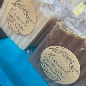 May include: Two bars of handmade goat milk soap wrapped in plastic. The soap on the left is labeled "Homemade Goat Milk Soap Volcano" and the soap on the right is labeled "Homemade Goat Milk Soap Rum  Rum".